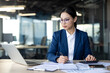 © Liubomir - Asian businesswoman working diligently in office setting, focused on laptop and paperwork, using calculator. Represents dedication, focus, and professionalism in workplace environment.
