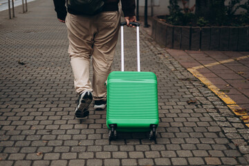 Unrecognizable man pulling green suitcase outdoors. Rear view. Copy space. Man walking on the street with travel bag.