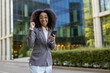 © Liubomir - African American business woman standing outside modern office building holding phone looking excited and confident. Expression of success, professional achievement, and positive energy in business
