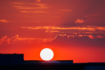 Stunning fiery sunset scene with vibrant red sky glowing sun setting behind city buildings urban silhouettes, creating warm dramatic atmosphere.  Tranquil horizon dusk cloudscape