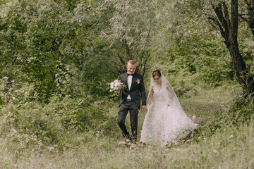 Wall Mural - A bride and groom are walking through a forest, holding hands. The bride is wearing a white dress and the groom is wearing a black suit. The scene is peaceful and romantic