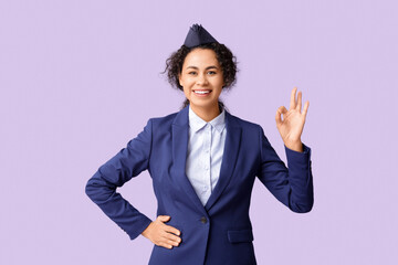 Wall Mural - Beautiful African-American stewardess showing OK on lilac background