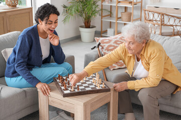 Wall Mural - Young African-American female medical worker playing chess with elderly woman in nursing home