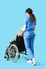 Wall Mural - Young African-American female medical worker with elderly woman on wheelchair against blue background