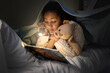 © Pixel-Shot - Little African-American girl with toy bear and flashlight reading book in bedroom at night