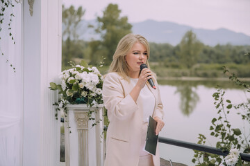 Wall Mural - A woman is standing on a balcony with a microphone, speaking into it. She is wearing a white jacket and is holding a clipboard. The scene takes place near a body of water