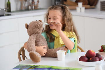 Sticker - Cute little girl with spoon feeding teddy bear yogurt in kitchen