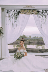 Poster - A woman in a white dress sits in front of a white archway. She is holding a bouquet of flowers