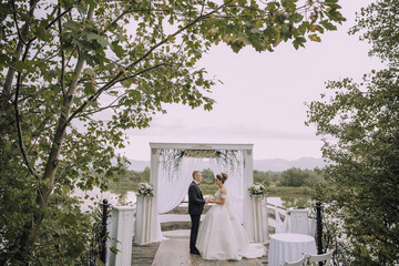 Wall Mural - A bride and groom stand under a white archway on a pier. The bride is wearing a white dress and the groom is wearing a black suit. The scene is serene and romantic