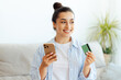 © Kateryna - E-banking concept. Cheerful pretty brunette woman, sits on a sofa in a living room, holds smartphone and credit card for online payment in a mobile app, making online shopping, looking away, smiling