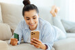 © Kateryna - Positive attractive caucasian young woman dressed in casual wear, lying on a sofa happily making an online purchase with a smartphone and credit card, order online delivery, pay bills, smiles