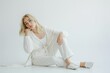 © miss irine - Blonde woman in white clothes sitting on white floor. Young adult female wears stylish outfit, elegant make-up, and has beautiful hair. Model sits alone, relaxed, and attractive on white background.