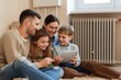 © miss irine - Happy family with tablet near heating radiator at home. Adult, children are comfortable, relaxed. Female, male parents are smiling with their kids. Central heating system provides warmth, comfort.