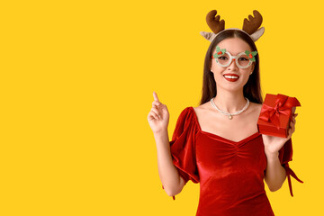 Canvas Print - Beautiful young Asian woman in Christmas deer horns and glasses with gift box on yellow background