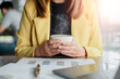 © Satori Studio - A woman in a yellow blazer holds a steaming coffee cup at a desk with business documents and a laptop in a sunlit office.