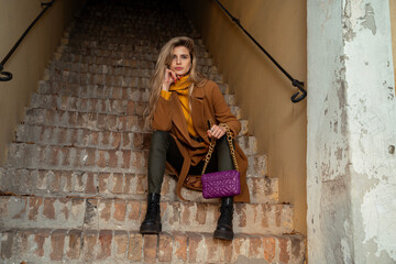 Attractive fit blonde girl in fall fashion clothes with bag, boots and coat sitting on rustic steps 