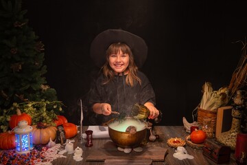 Halloween, smiling little cute girl dressed as witch in witch's hat brews a glowing magic potion in cauldron on coals. Magic wand, сandles, pumpkins, ingridients, fog smoke, magic, mystical atmosphere