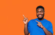 © Iryna - A cheerful young man in a blue shirt playfully gestures pointing towards empty place an orange background with a confident smile