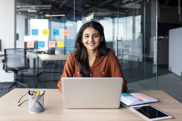 Wall Mural - Confident woman sitting at desk in contemporary office setting, using laptop for work. Background shows glass wall with colorful sticky notes. Scene conveys productivity, professionalism