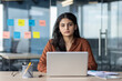 © Liubomir - Young professional woman sits at desk with laptop and open book, focusing on work in modern office. Colorful sticky notes on glass wall suggest brainstorming, planning, teamwork.