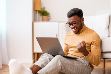 Canvas Print - Big Luck. Excited Black Guy Using Laptop Shaking Fists Celebrating Success Sitting On Floor At Home. Selective Focus