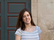 © BeatrizHerrera - A young woman with long hair stands confidently outside a historical building's entrance, wearing a casual striped top and expressing joy, enjoying a sunny day.