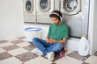 © Prostock-studio - A young individual sits on the floor of a laundromat, wearing headphones and looking at a phone. They are relaxed and engaged while laundry machines operate in the background.