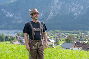 Wall Mural - An elderly farmer in national dress, leather shorts and a cap with a feather stands against a backdrop of mountains and green grass.