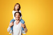 © Prostock-studio - Handsome dad carrying his lovely cheerful little daughter on shoulders, posing over yellow background, copy space