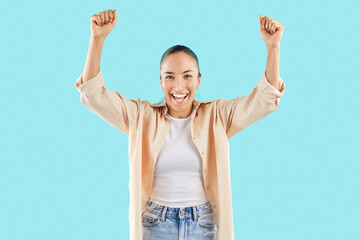 Portrait of a Caucasian young adult woman celebrating success and achievement, exclaiming yes and raising hands in accomplishment. She is happy and joyful, set against a blue background.