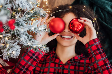 Close-up funny portrait of young happy African woman in plaid pajamas with red Christmas balls. Cute black girl is laughing and decorating Christmas tree at home.