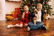 © Dasha Petrenko - Happy sibling in fancy clothes are sitting under the Christmas tree. Cute brother and sister are smiling and holding New Year gifts in the living room.