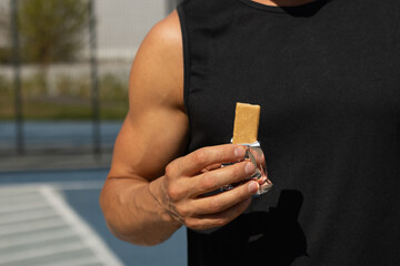 Sports man eating energy bar. Muscular athlete snacks on protein bar while resting after workout at stadium. Healthy protein snack or energy bar, fitness dieting food for sporty lifestyle.