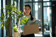 © Alexandra - New employee starting new job holding cardboard box with belongings and plant smiling in modern office