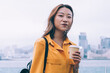 © BullRun - Half length portrait of attractive female in trendy apparel posing at Hong Kong landscape with coffee to go and digital tablet in hands, charming Chinese student standing at urban setting near river