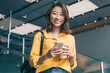 © BullRun - Half length portrait of pretty hipster girl with cellphone device for blogging standing indoors and rejoicing, joyful Asian woman feeling happiness during online communication via smartphone
