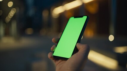 Side view of person holding smartphone vertically with one hand. Chroma key visible on large display. Pressing on touch screen with one finger. Green screen. Evening in city. Bright lights.