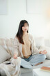 © Jirapaporn - An Asian woman meditating on a sofa.
