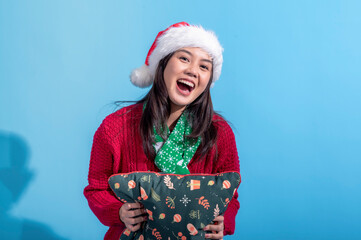 An Asian woman dressed in a red sweater and Santa hat smiles warmly as she hugs a Christmas-themed pillow decorated with holiday patterns. She stands against a light blue background