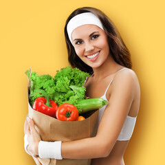 Happy smiling young beautiful brunette woman in fitness wear holding grocery shopping bag with vegetarian raw food, isolated orange yellow studio background. Healthy eating and dieting concept. Square