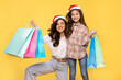 © Home-stock - Young woman and her child girl both in Santa hats, holding shopping package bags isolated on yellow background. Xmas winter shopping concept