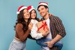 © Home-stock - Happy Xmas and New Year holidays. Positive European family with child girl holding Christmas gift, posing over blue background and smiling at camera, wearing Santa hats