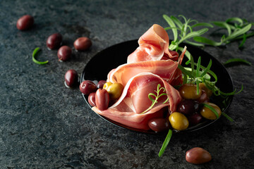 Poster - Italian prosciutto crudo with olives and rosemary on a black stone table.