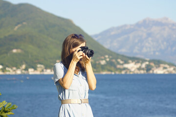 Female photographer during outdoor photoshoot: woman taking photo on the coast holding professional camera to her face. Girl with DSLR camera on the background of nature in Kotor Bay, Montenegro.