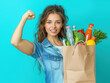 © Petzhy - A strong woman in denim proudly flexes her bicep while holding grocery bag filled with fresh produce and drinks, showcasing her healthy lifestyle and confidence