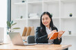 © PRIME STOCK LAB - professional woman is sitting at desk, smiling while holding notebook and looking at laptop. bright office space features plants and organized shelves, creating productive atmosphere.