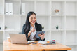 © kenchiro168 - young woman in black suit smiles while using tablet at her desk, surrounded by office supplies and laptop. Her cheerful demeanor reflects productive work environment.