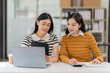 © ArLawKa - Two Asian women work online with laptop and smartphone. A joyful moment captured as two female colleagues laugh together during a productive meeting in the office.