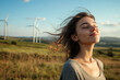 © Martí Rosselló - Young Woman Enjoying Fresh Air in Wind Farm Landscape, A young woman stands outdoors with her eyes closed, enjoying the breeze in a serene landscape with wind turbines in the background.