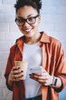 © BullRun - Half length portrait of happy female blogger holding takeaway cup with caffeine beverage and smartphone gadget in hands, cheerful teenager in eyeglasses smiling at camera while waiting sms answer